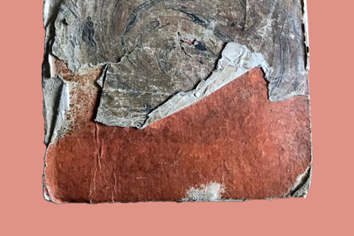 Close-up of a weathered, layered paper fragment with peeling textures and earthy red and gray hues against a muted pink background.